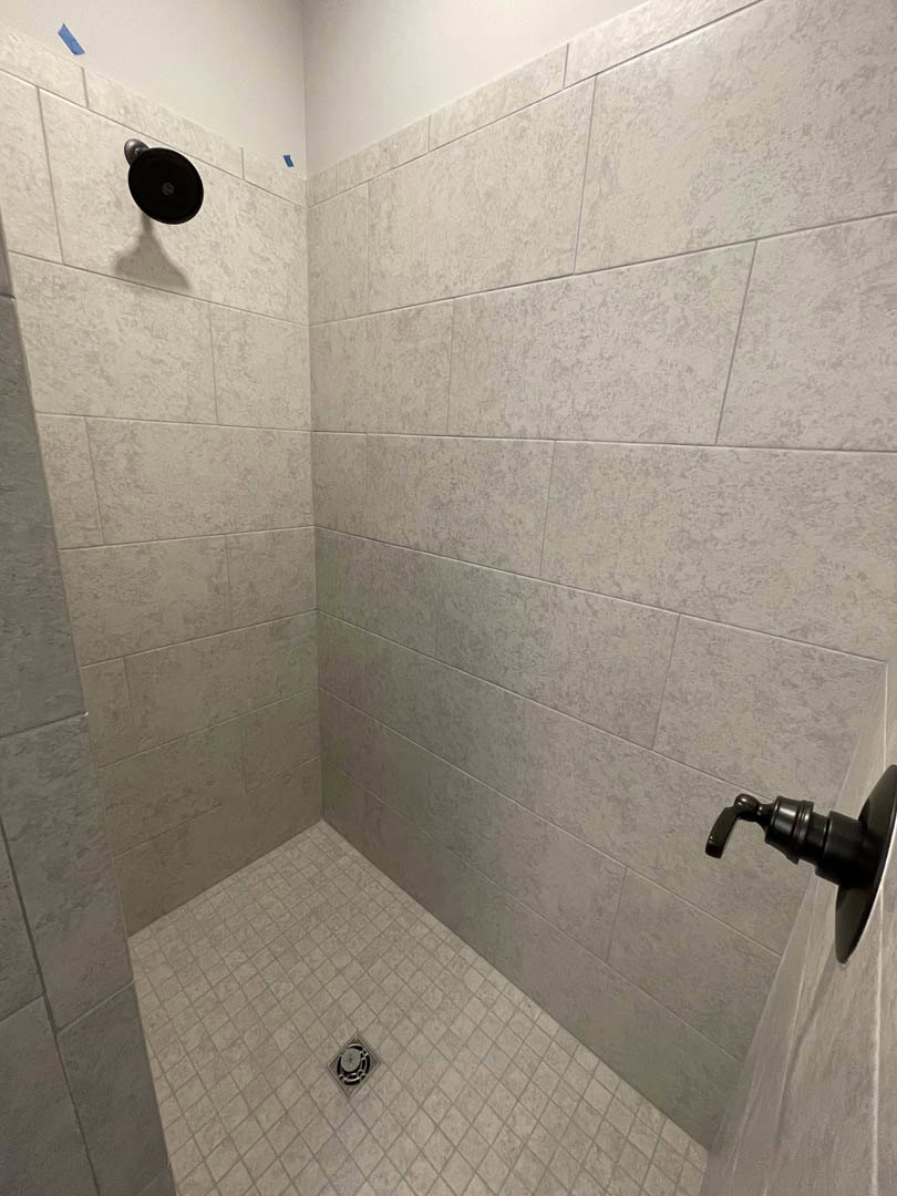 Shower with black shower head and handle, black soap dispenser mounted on tiled wall, circular vent, tiled shower floor with drain