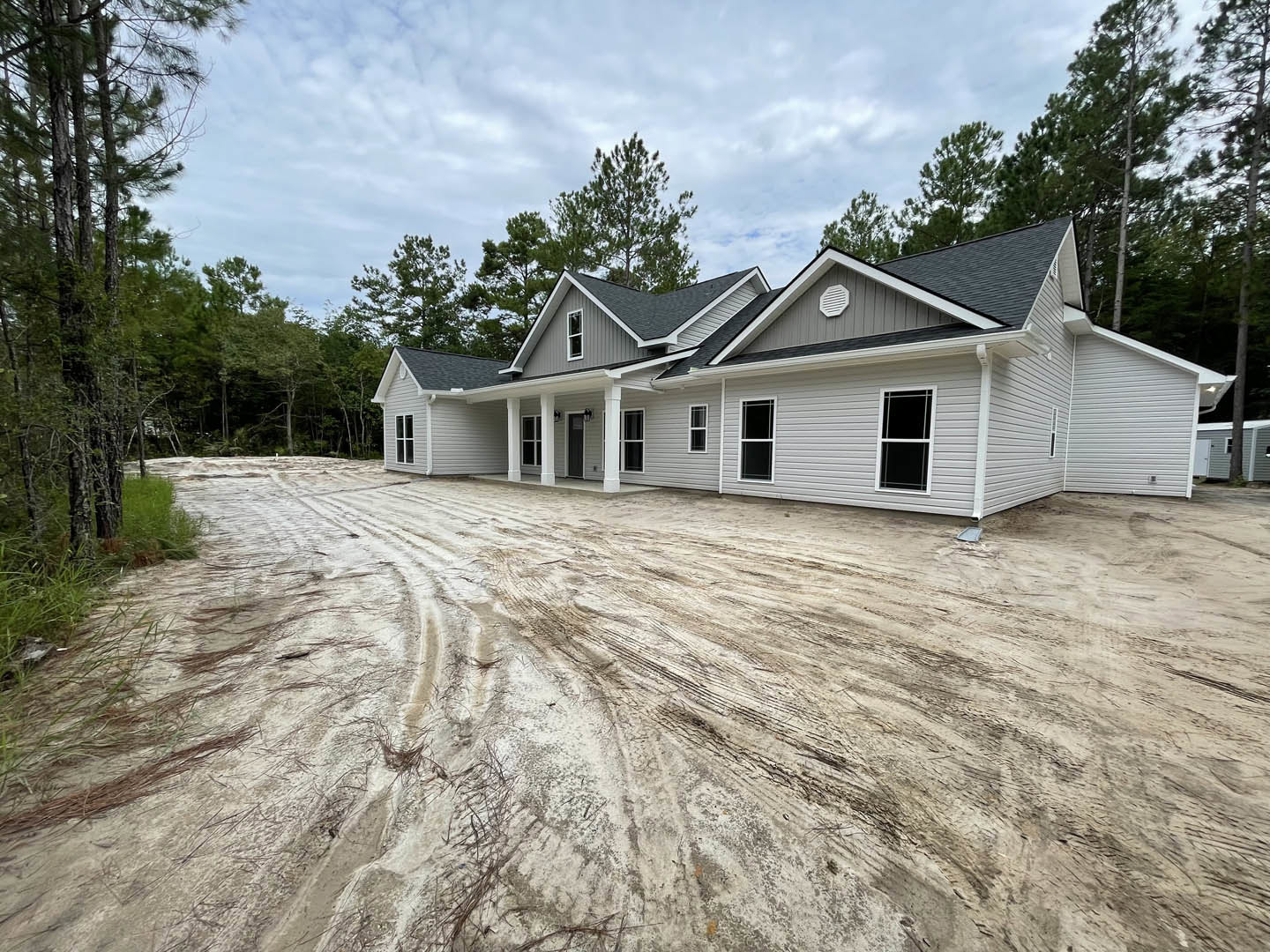 Partially built house with exposed roof and window, surrounded by dirt ground and tire tracks, black screened window visible, dense trees in background under cloudy sky