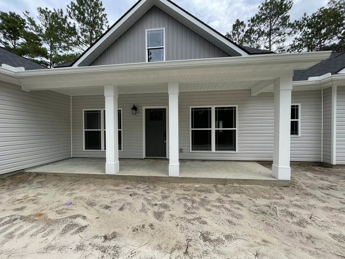 Two-story house with white siding, black front door, white-framed windows, covered porch, and concrete driveway on sandy ground with scattered debris