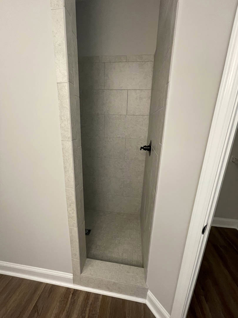 Walk-in shower with matte black faucet, white plaster walls, and light wood flooring