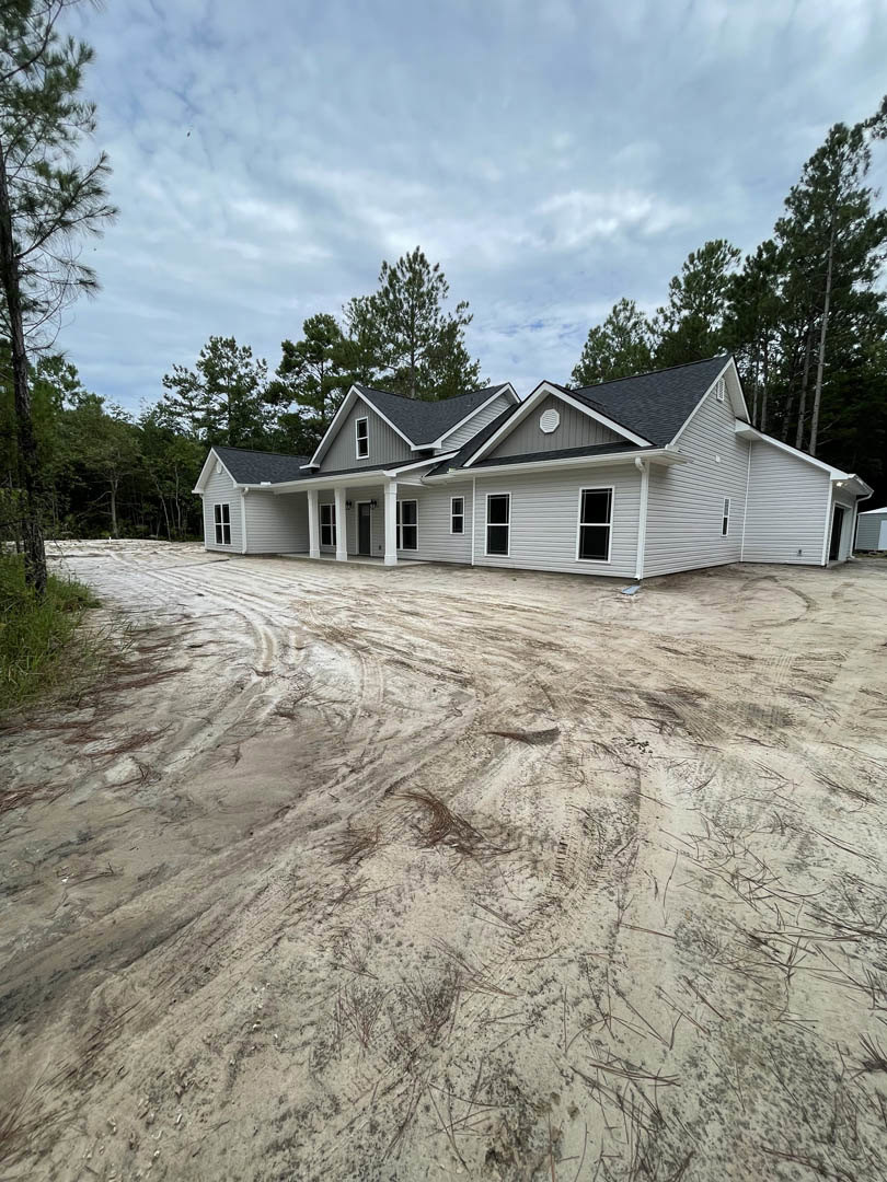 Grey-roofed house surrounded by trees, dirt road with tire tracks leading to the property, patches of snow on the ground, cloudy sky overhead