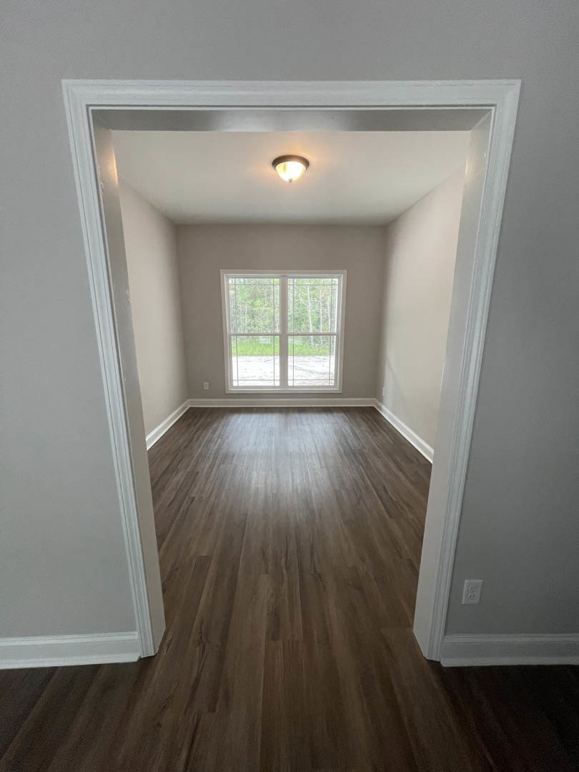 Wood flooring with white trim, large window with white frame, plaster walls, ceiling light fixture, natural light illuminating interior space.