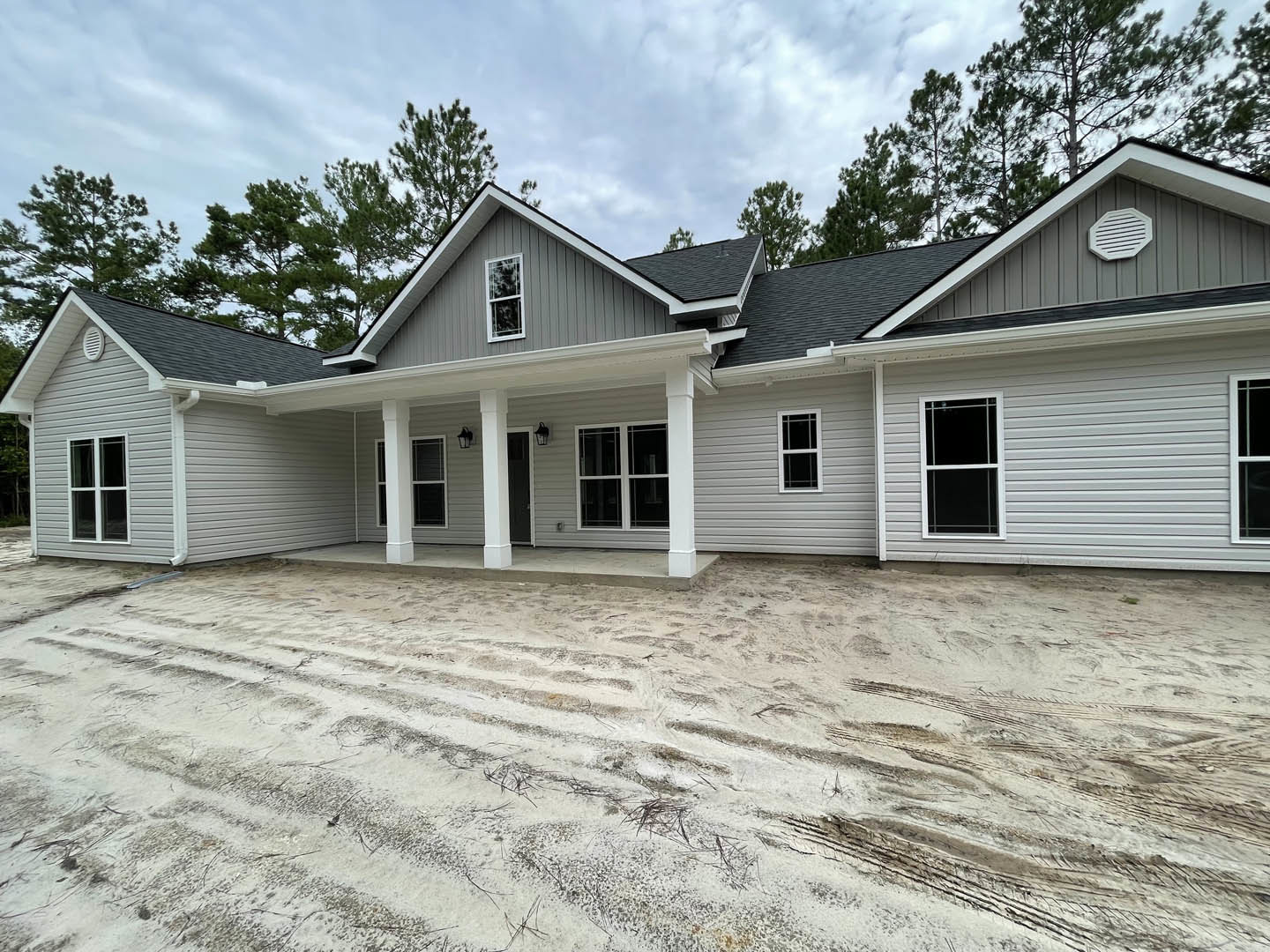 Two-story house with white porch, white door, and white window frames, dirt driveway in front, black screened window, trees and cloudy sky in background