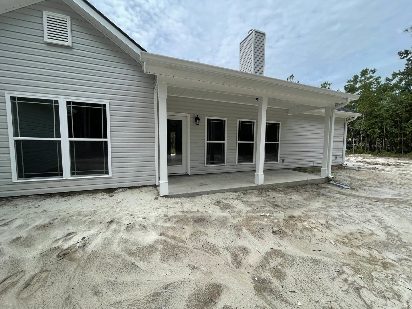 Grey siding home with white-framed windows, white vent, covered porch, chimney, sandy backyard, and partly cloudy sky