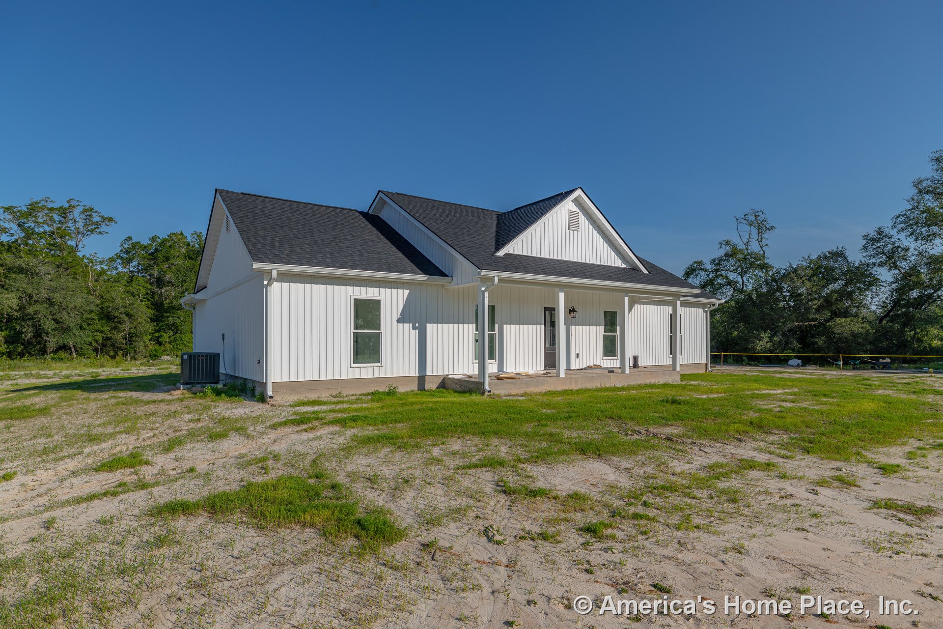 White vertical board and batten siding with black shingle roof, covered front porch supported by columns, multiple rectangular windows, exterior wall-mounted light fixture, and