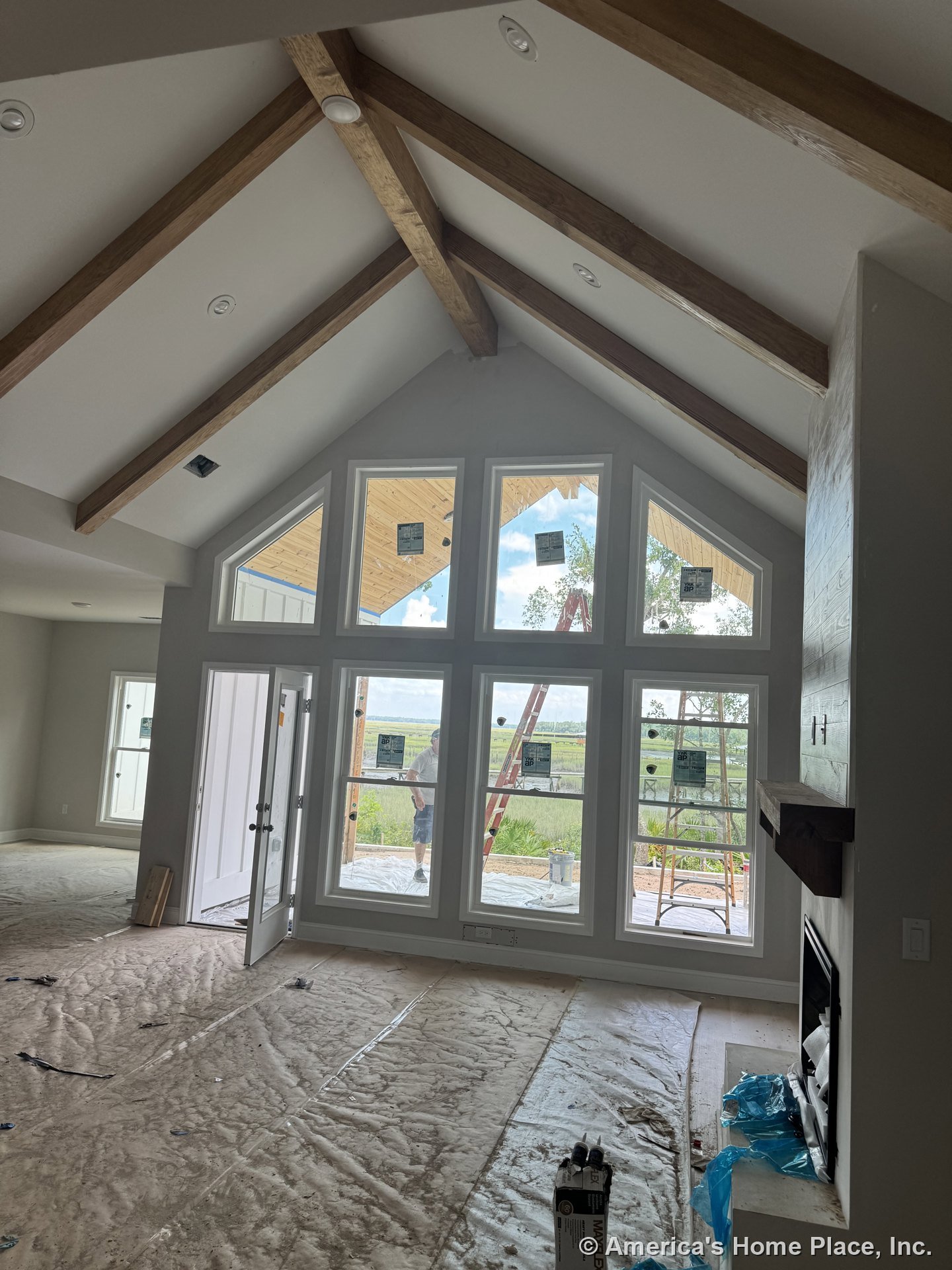 Vaulted living room ceiling with exposed wood beams, floor-to-ceiling windows framed in white trim, built-in fireplace with wood mantel, recessed ceiling lights, and covered porch