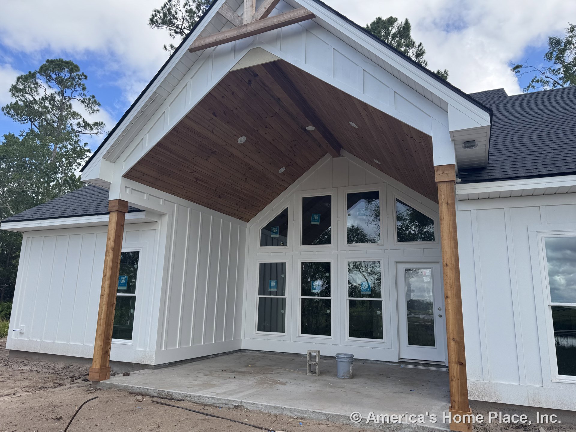 Covered front porch with vaulted wood ceiling, wooden support columns, board and batten siding, large grid windows, white exterior trim, concrete floor, and permanent lighting at