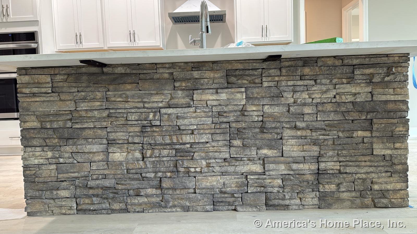 Stacked stone veneer kitchen island with white quartz countertop, built-in sink, modern faucet, white shaker cabinets, stainless steel vent hood, and light wood flooring.