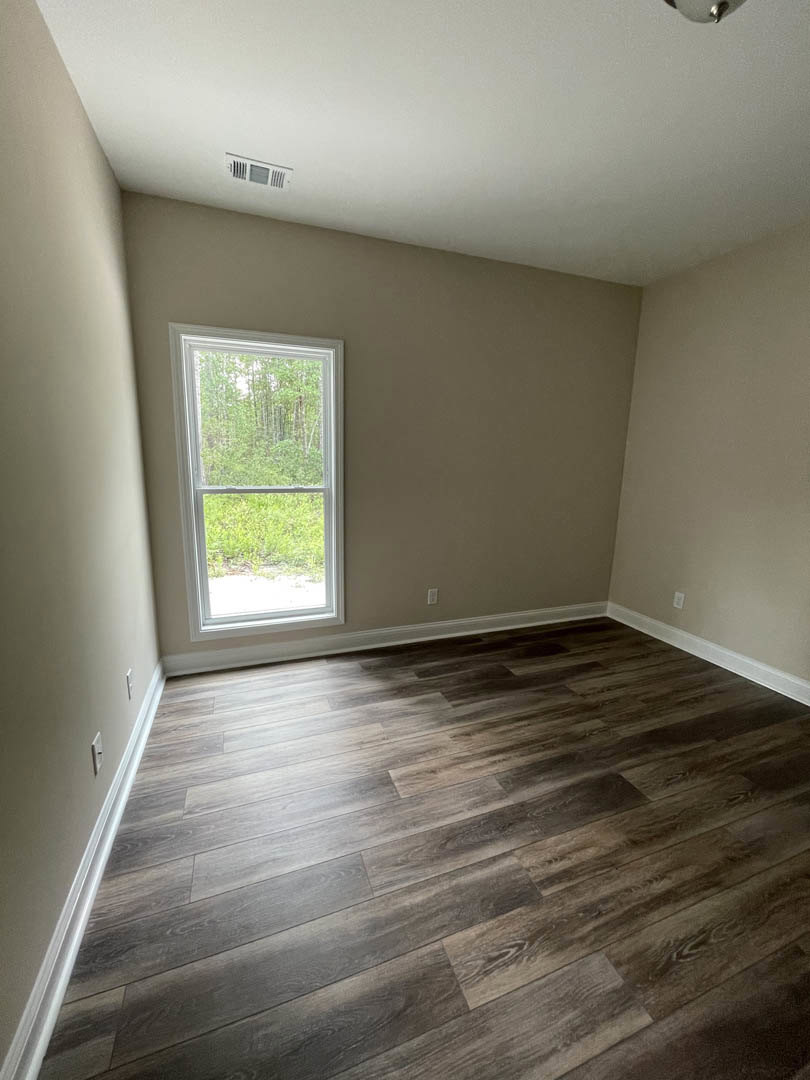 Sunlit room featuring a large window overlooking trees, smooth hardwood flooring, white plaster walls, and a white ceiling with an air vent.