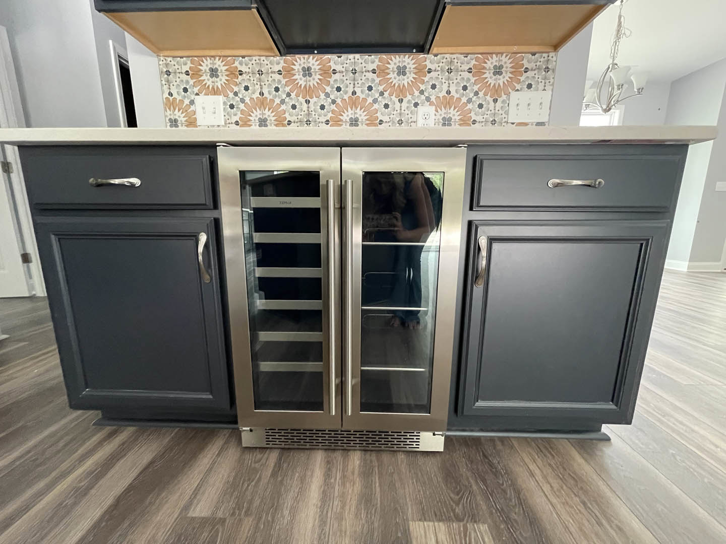 Stainless steel refrigerator with glass doors beside dark cabinetry and drawers, light stone countertop, hardwood flooring, woman standing in front of refrigerator