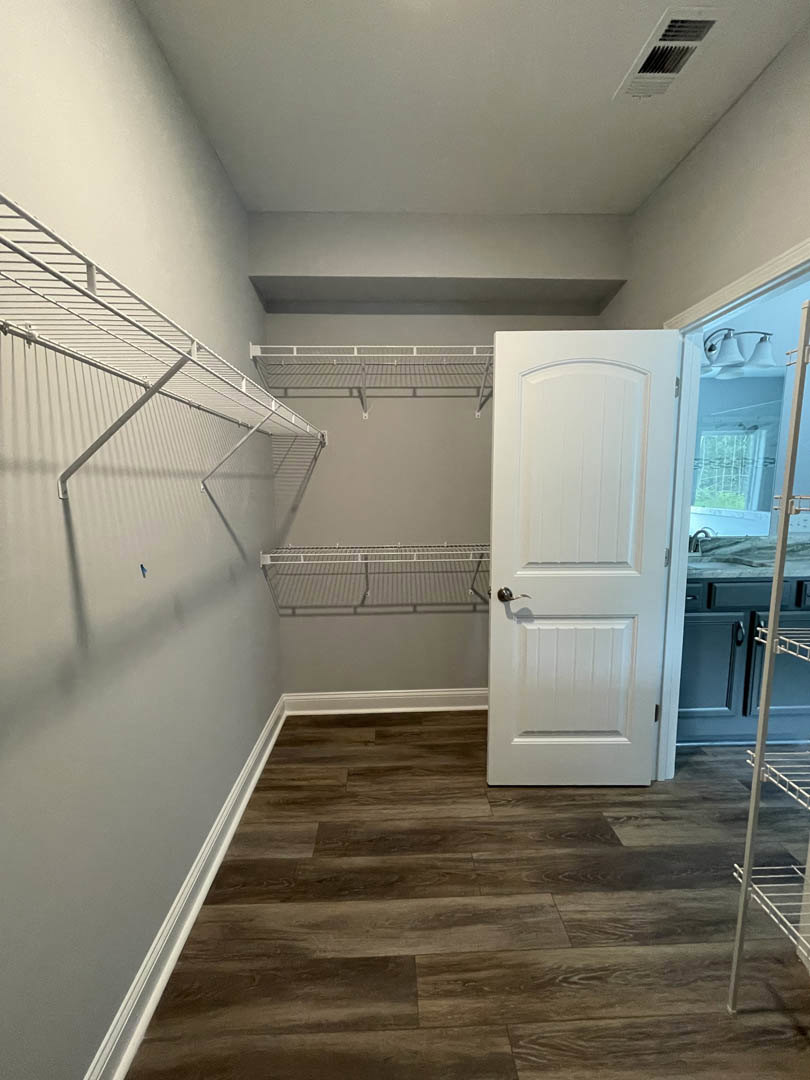Open white closet door revealing wood flooring, white shelving, and plaster walls; metal grid detail visible, lamp blurred in background.