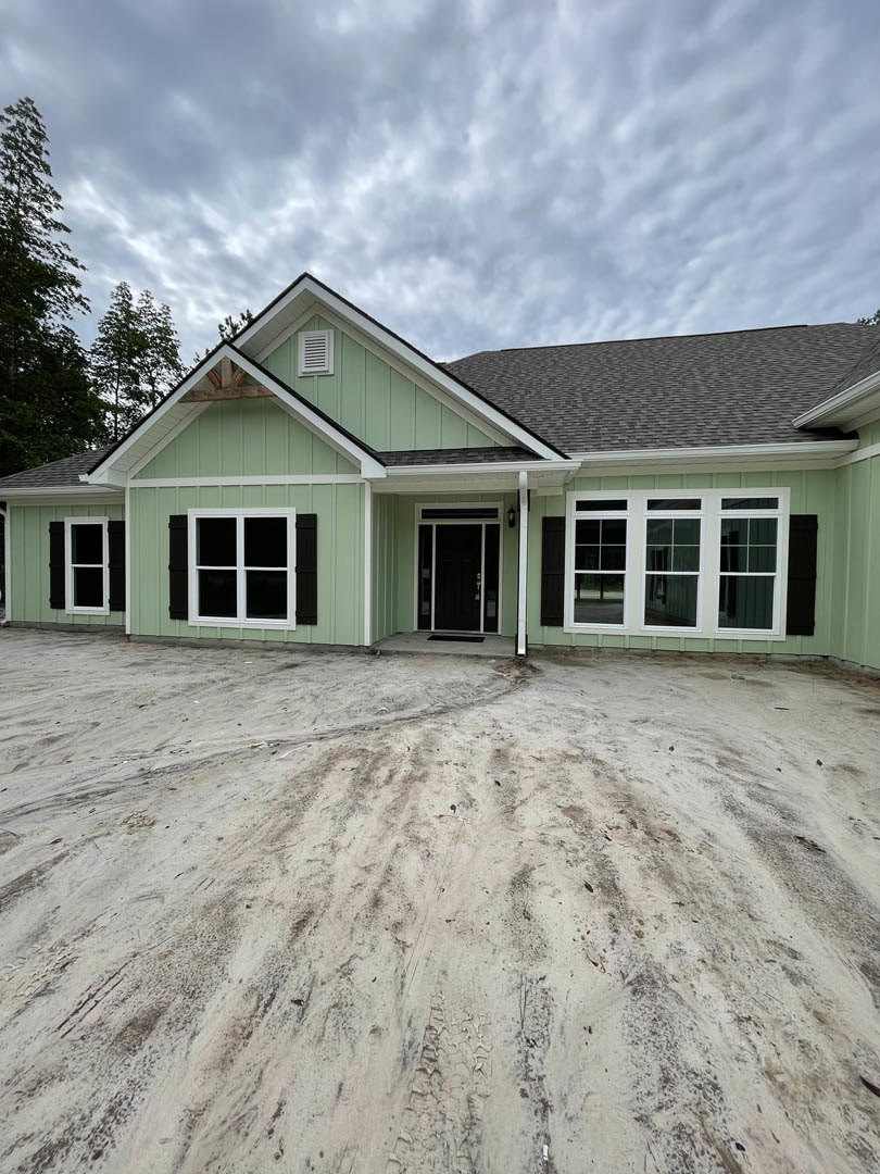 Green house with black door, white-framed window, glass panel door, tire tracks in sandy ground, dirt and trees behind, cloudy sky above