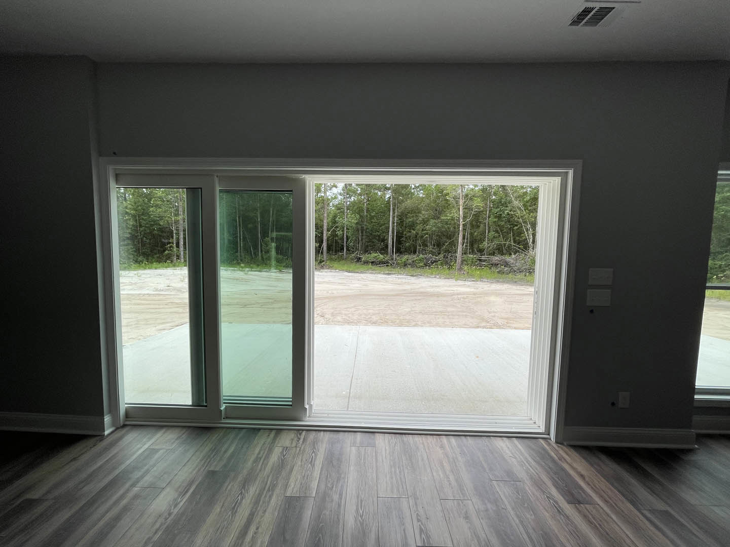 Wood laminate flooring in a bright room with a large sliding glass door, trees and a road visible outside, ceiling vent overhead.