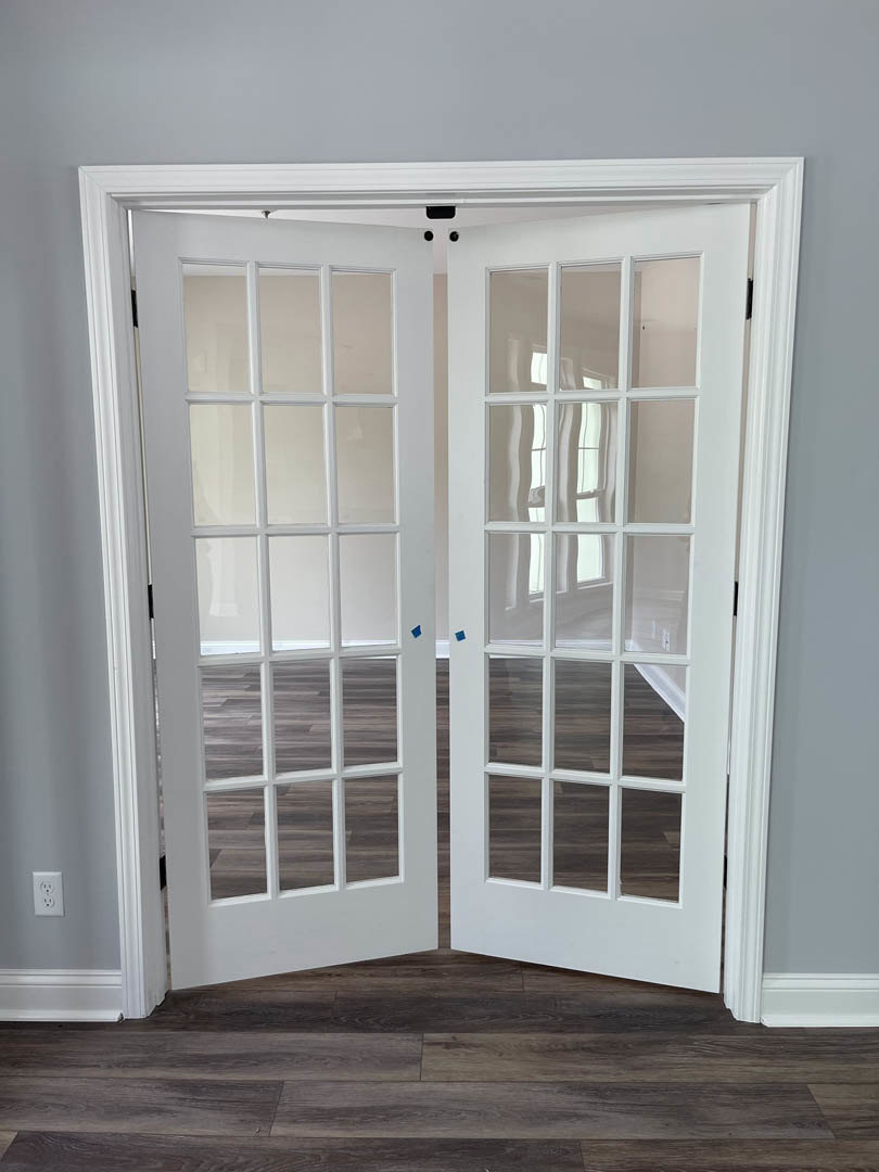 White double doors with glass panes set in a room with wood flooring, white walls, and a window visible nearby