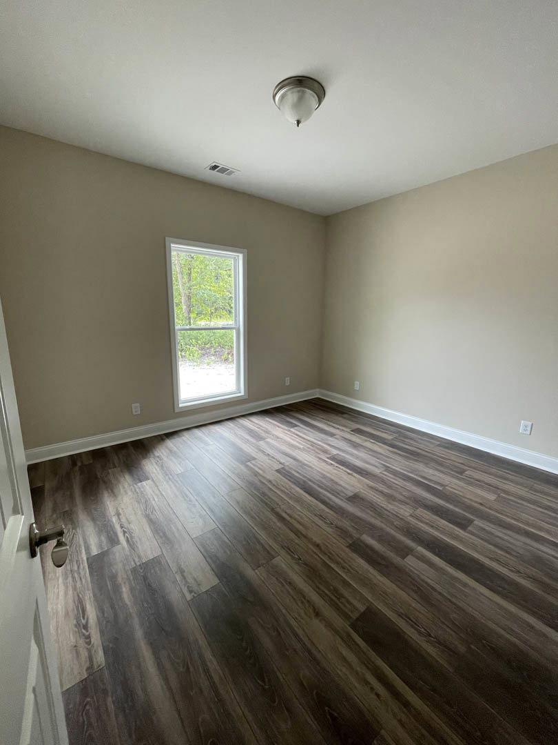 Room with hardwood floor, white walls, ceiling light fixture, large window overlooking trees, white door frame