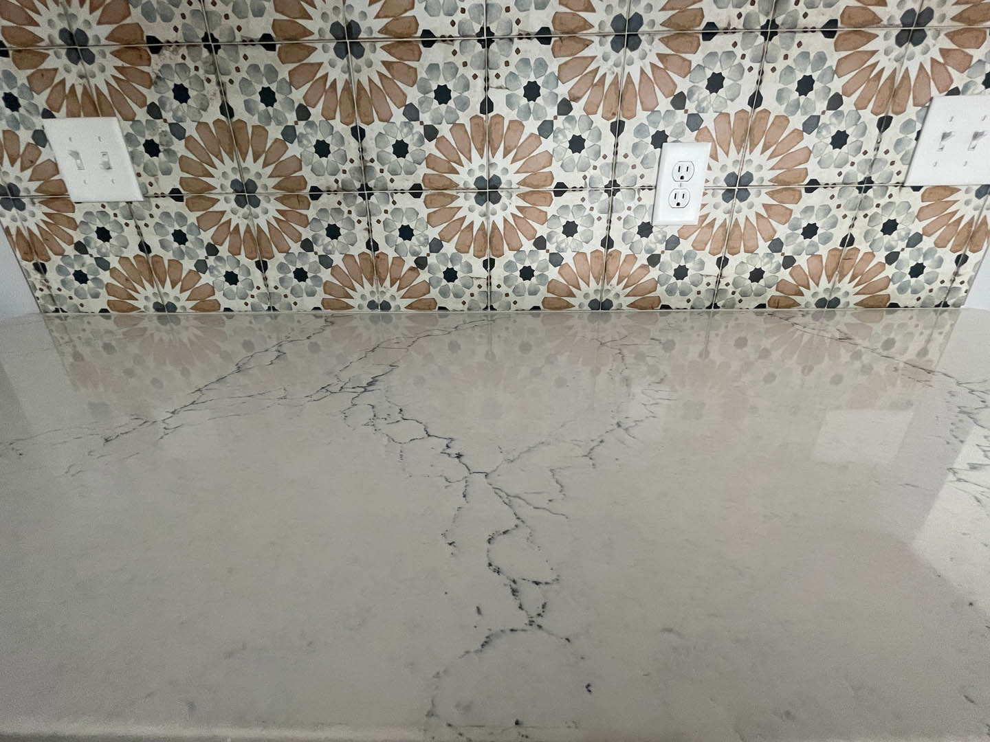 Marble countertop with patterned tile backsplash, white marble floor showing a visible crack, close-up of decorative tile, electrical outlet, and white light switch on wall