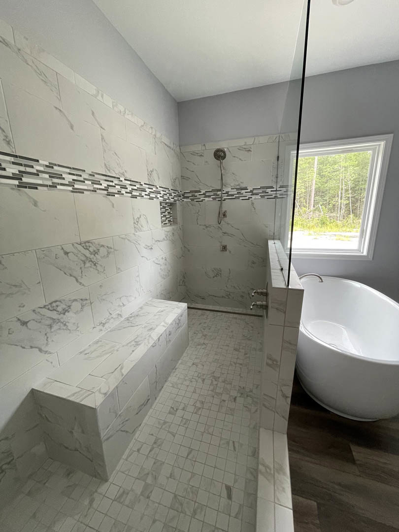 Bathroom featuring a freestanding white tub, glass-enclosed shower with marble tile walls, white tiled floor, built-in bench, large window overlooking trees, and modern plumbing