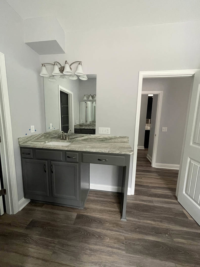 Bathroom featuring marble countertop, rectangular mirror above sink, white cabinetry with drawers, three-light fixture, wood flooring, white door with matching frame, open cabinet