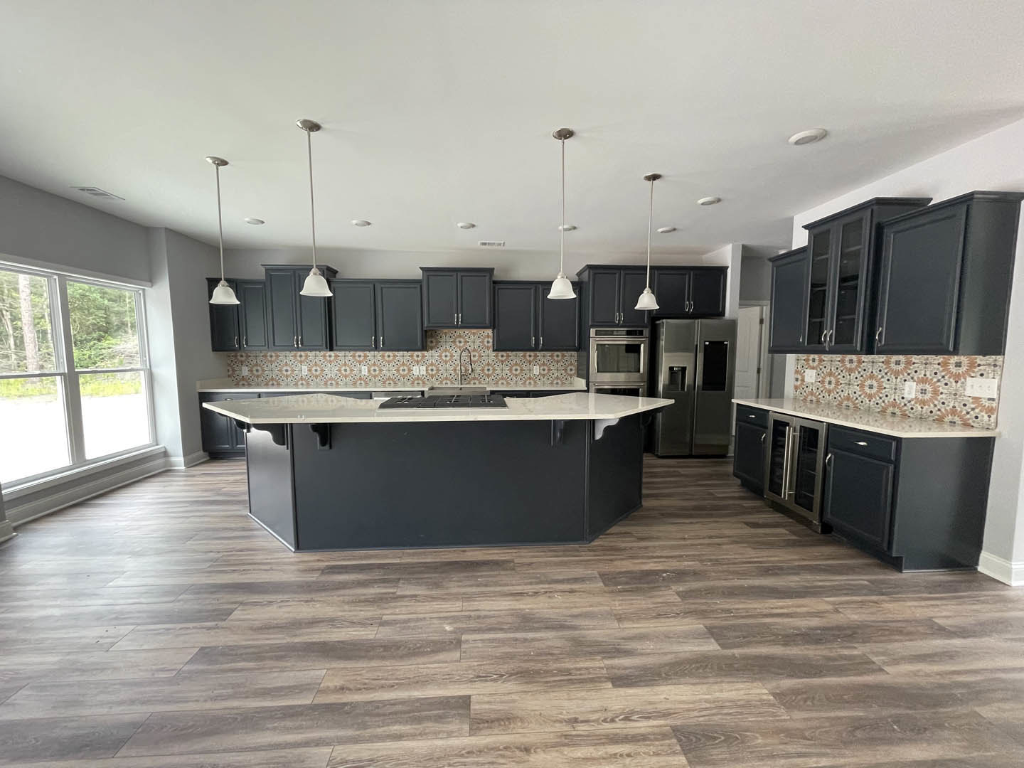 Spacious kitchen featuring a large white island with integrated sink, wood flooring, black cabinetry, stainless steel refrigerator and microwave, tile backsplash, and a window