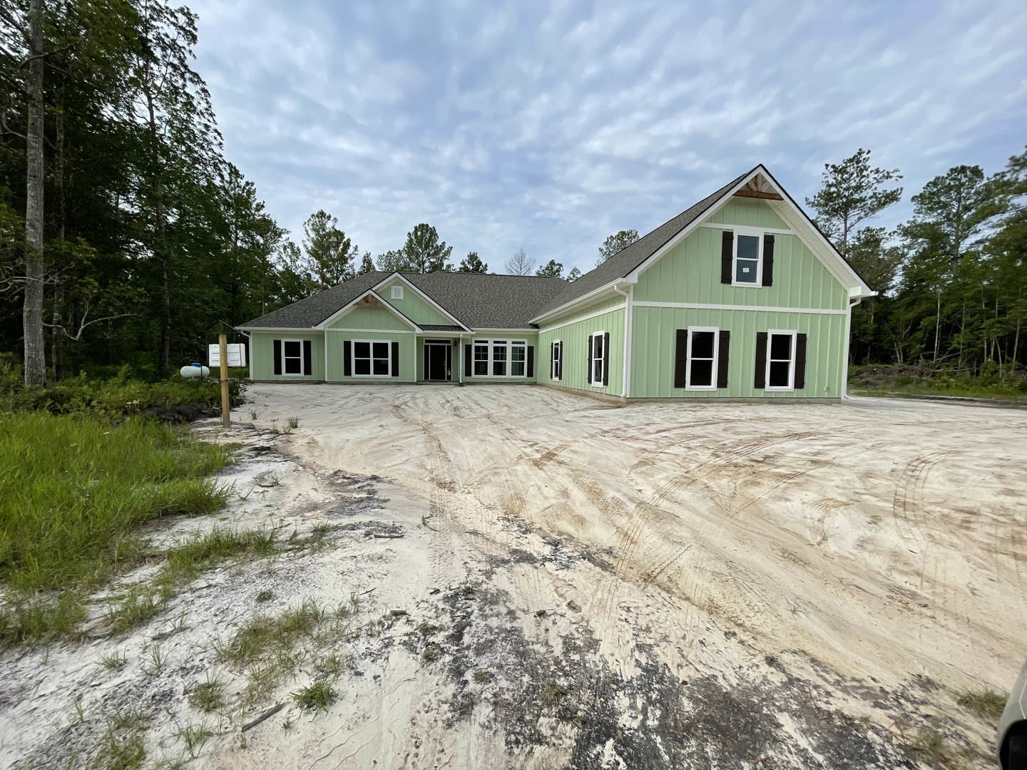 Single-story home with light siding, white-framed windows, dirt driveway, and mature trees in the background under a partly cloudy sky