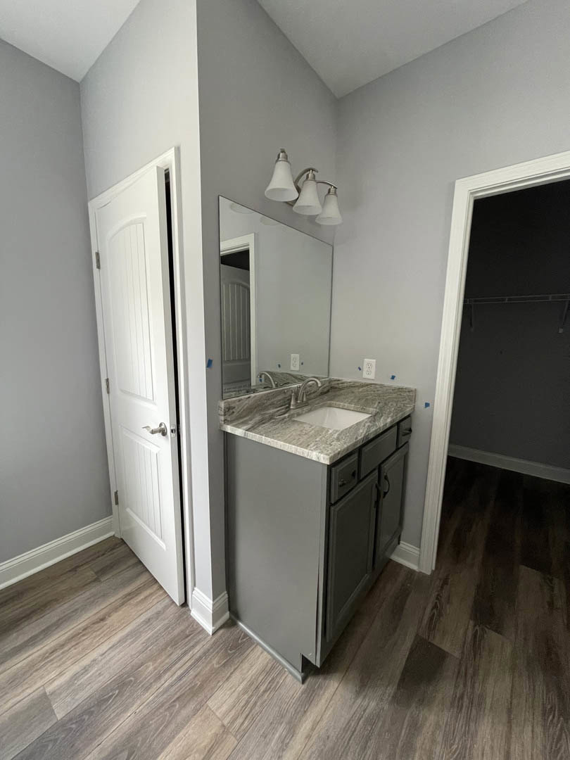 Bathroom with marble countertop, rectangular mirror above sink, three-light fixture, white cabinetry with silver handles, and white door adjacent to vanity