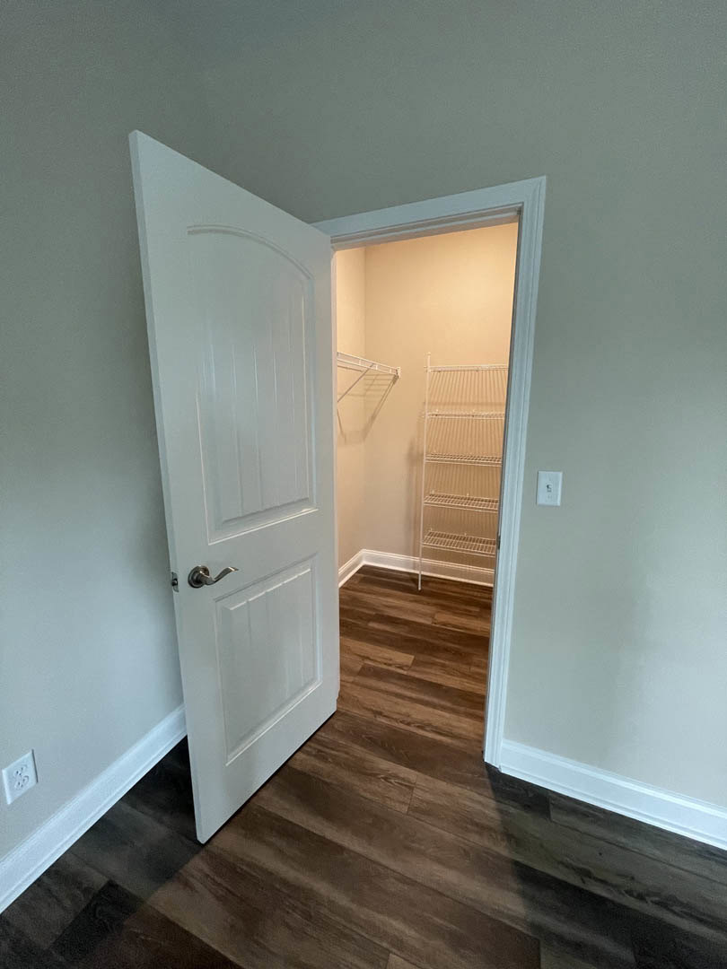 Open white door revealing empty closet with built-in white plywood shelves, plaster walls, and light wood flooring