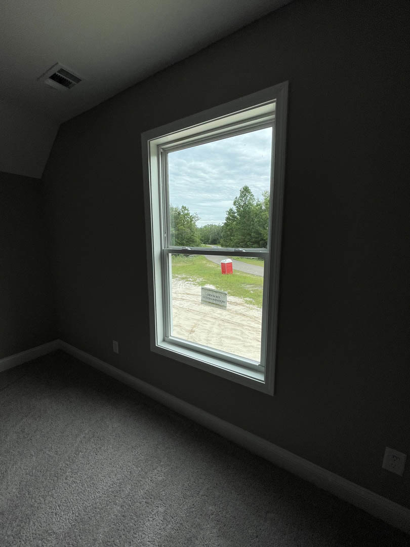 Large window set in a white wall, natural light streaming onto light wood floors, with a black and white sign visible outside and leafy trees beyond.