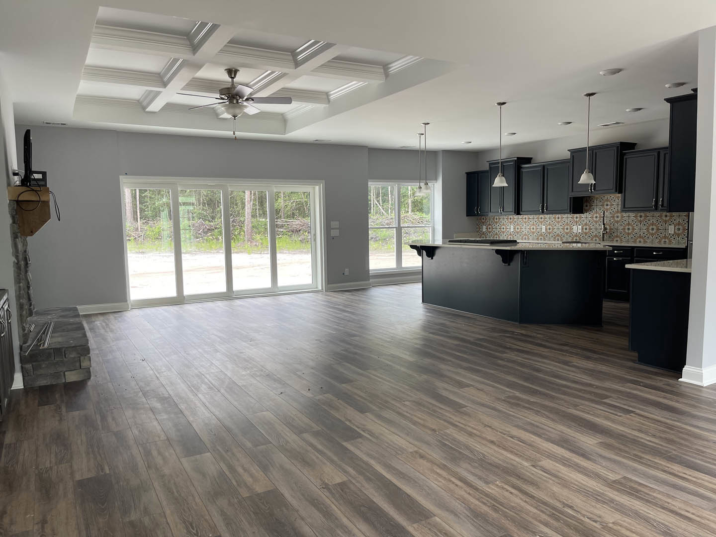Open kitchen and living room with hardwood flooring, black countertop, cabinetry, stone accent wall with metal bars, ceiling fan with light fixture, large windows and doors