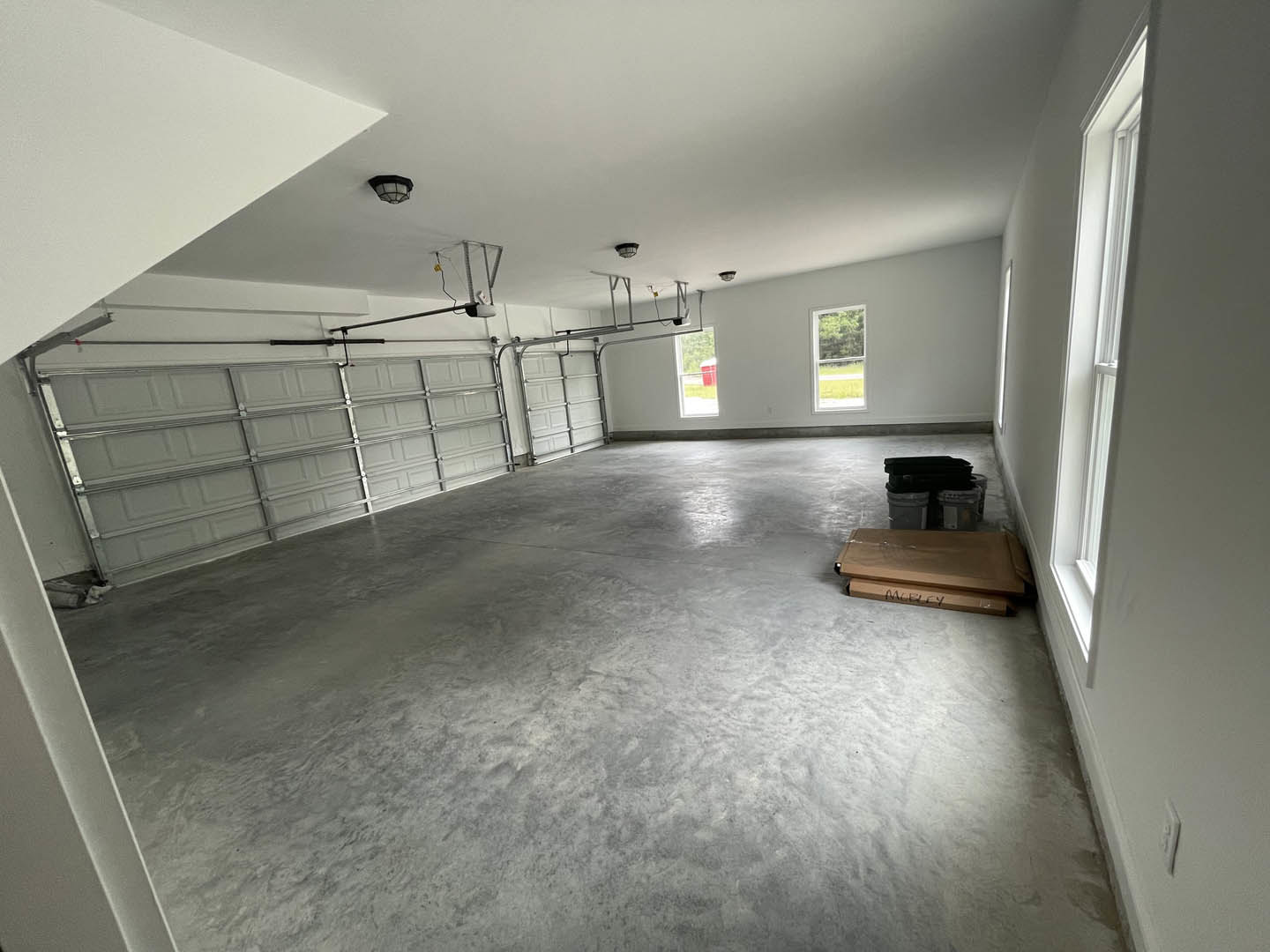 Concrete-floored garage with white plaster walls, large window overlooking fenced yard with trees, metal-barred garage door, brown storage box, and black object casting a shadow.