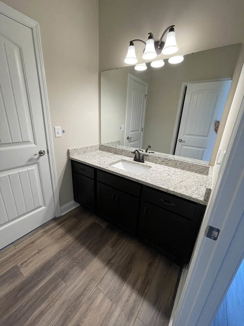 Marble countertop with undermount sink, large framed mirror, white door with silver handle, wood flooring, and chrome light fixture in a bathroom.