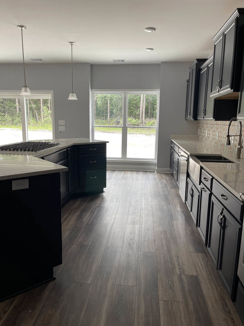 Kitchen with wide plank wood flooring, white cabinetry, black rectangular island with wood countertop, stainless steel sink, large windows overlooking trees, white ceiling with