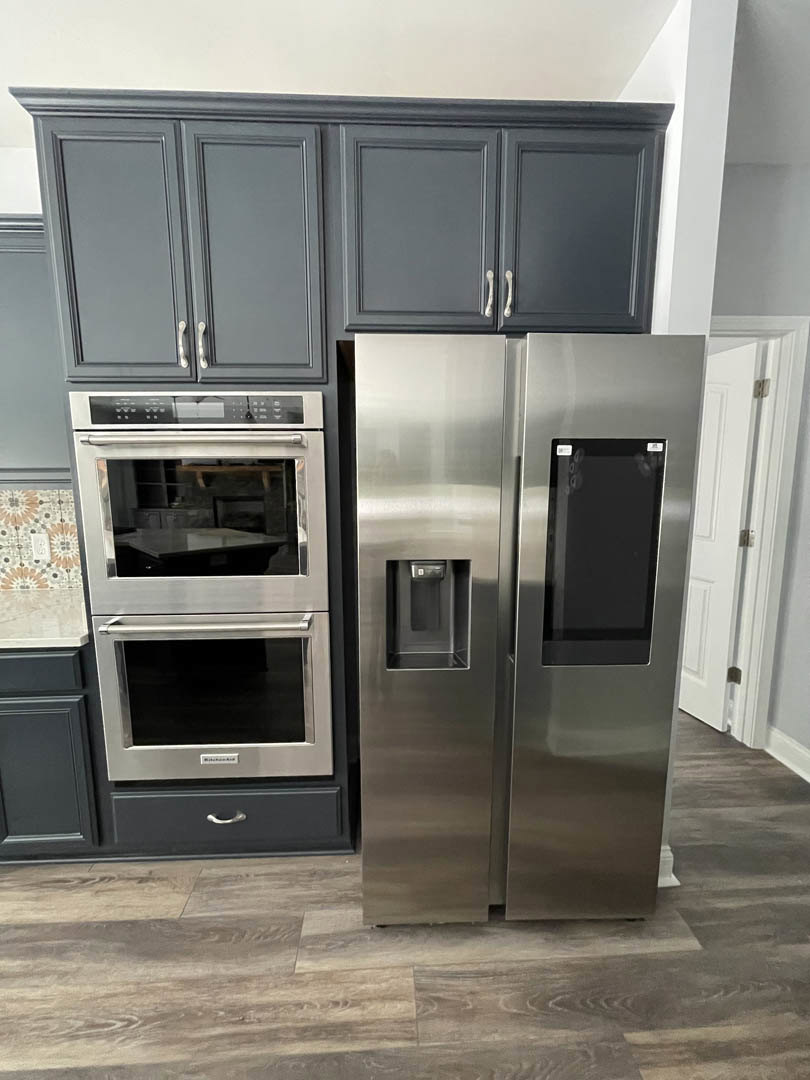 Modern kitchen featuring stainless steel refrigerator with black screen, built-in oven and microwave, white cabinetry, stone countertops, and a window-side dining table