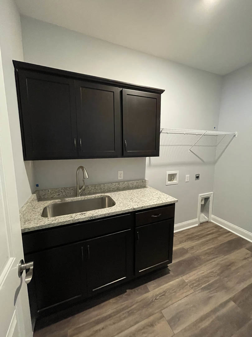 Black kitchen cabinets with brushed metal hardware, white tile backsplash, stainless steel sink and faucet, light-colored stone countertop, white electrical outlet, and wood