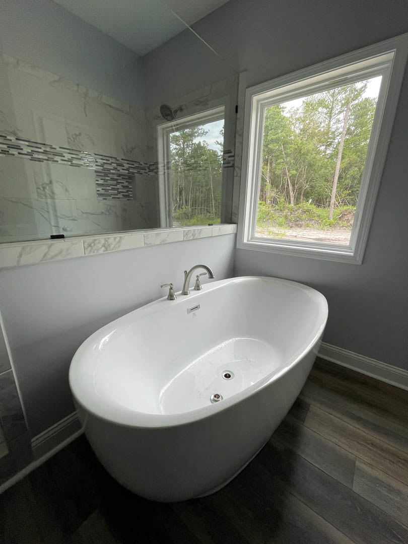 Freestanding white bathtub with chrome faucet beside a tiled wall, large window overlooking trees, bathroom interior with modern finishes