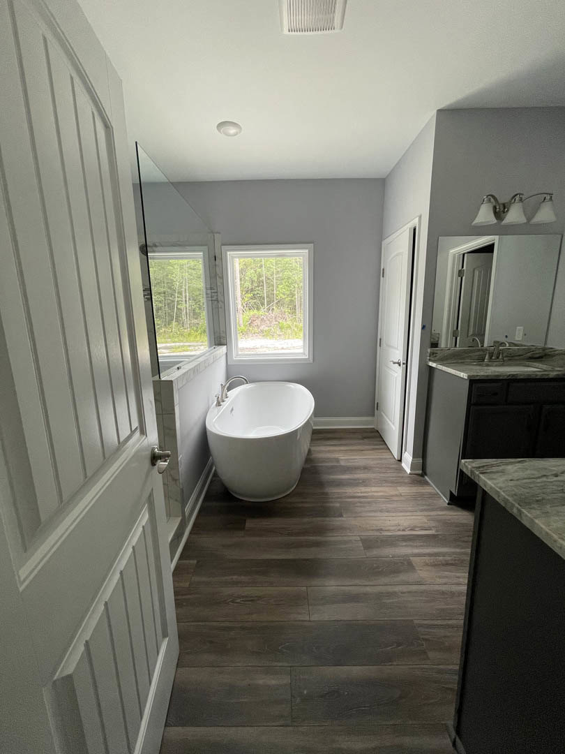 White freestanding bathtub on wood floor beside window with tree view, wall-mounted sink, cluster of modern lights, tile accents, and black board detail