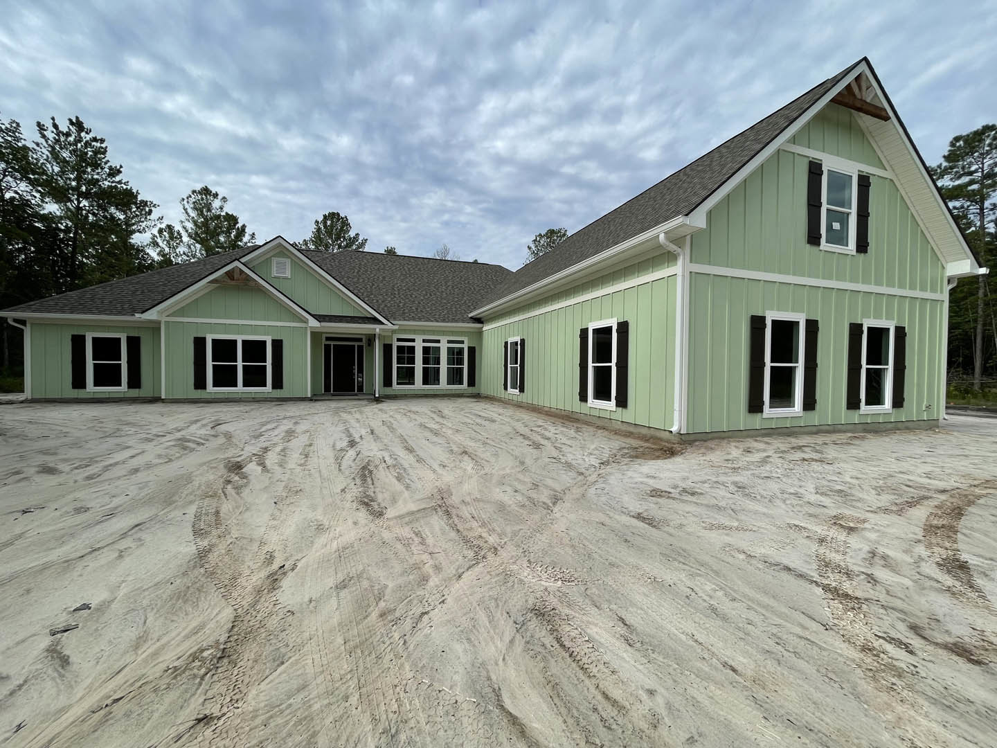 Modern home exterior with white-framed windows, dirt driveway showing tire tracks, cloudy sky overhead, and sparse landscaping with trees in the background
