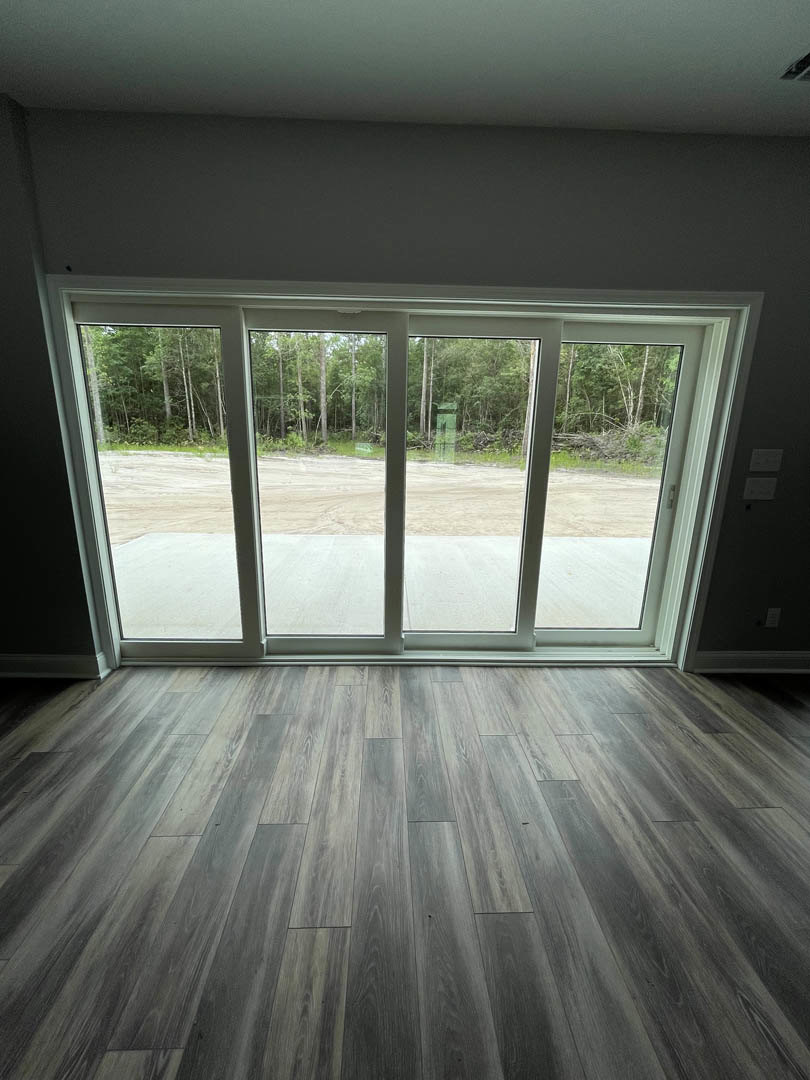 Wood flooring and sliding glass door opening to forest view under cloudy sky, natural light filling modern interior space.