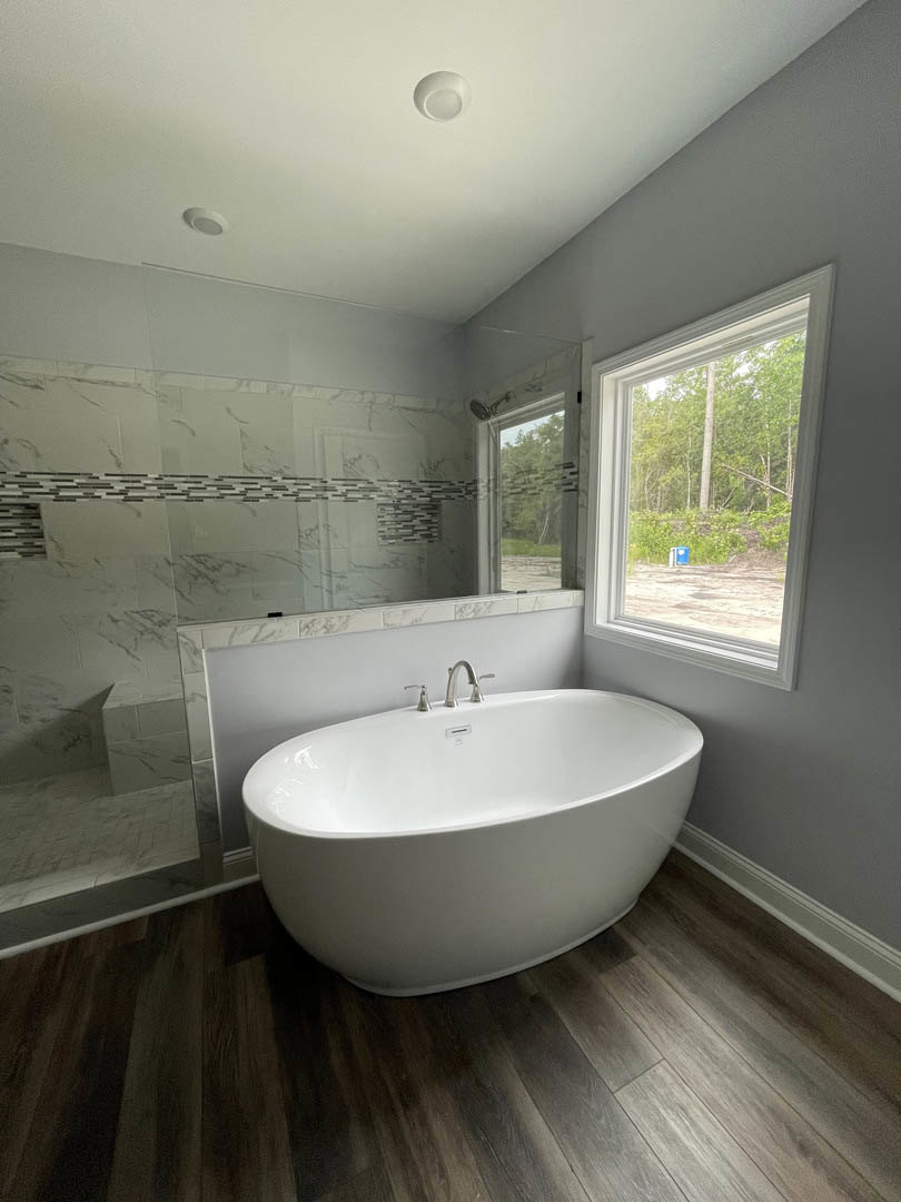 Freestanding white bathtub with chrome faucet on wood floor, large window with view of trees, white tile walls, modern bathroom design
