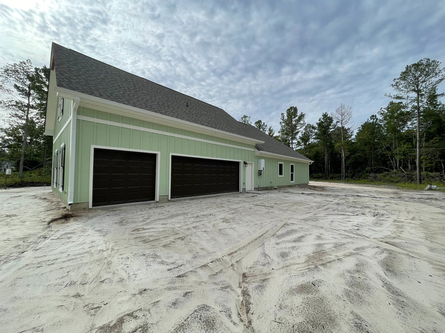 Black garage door with white trim on green exterior wall, snow-covered driveway with tire tracks, tall trees in wooded background