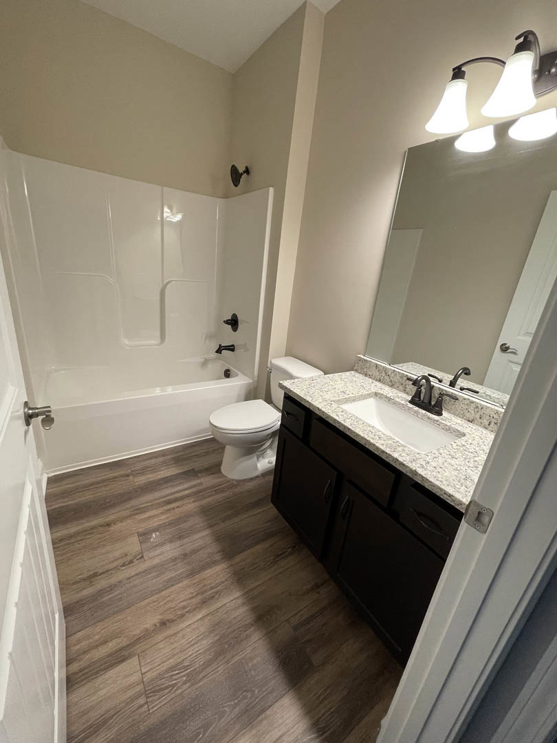 Bathroom with wood flooring, white sink featuring a black faucet, toilet beside a white bathtub, and a three-light fixture mounted above.