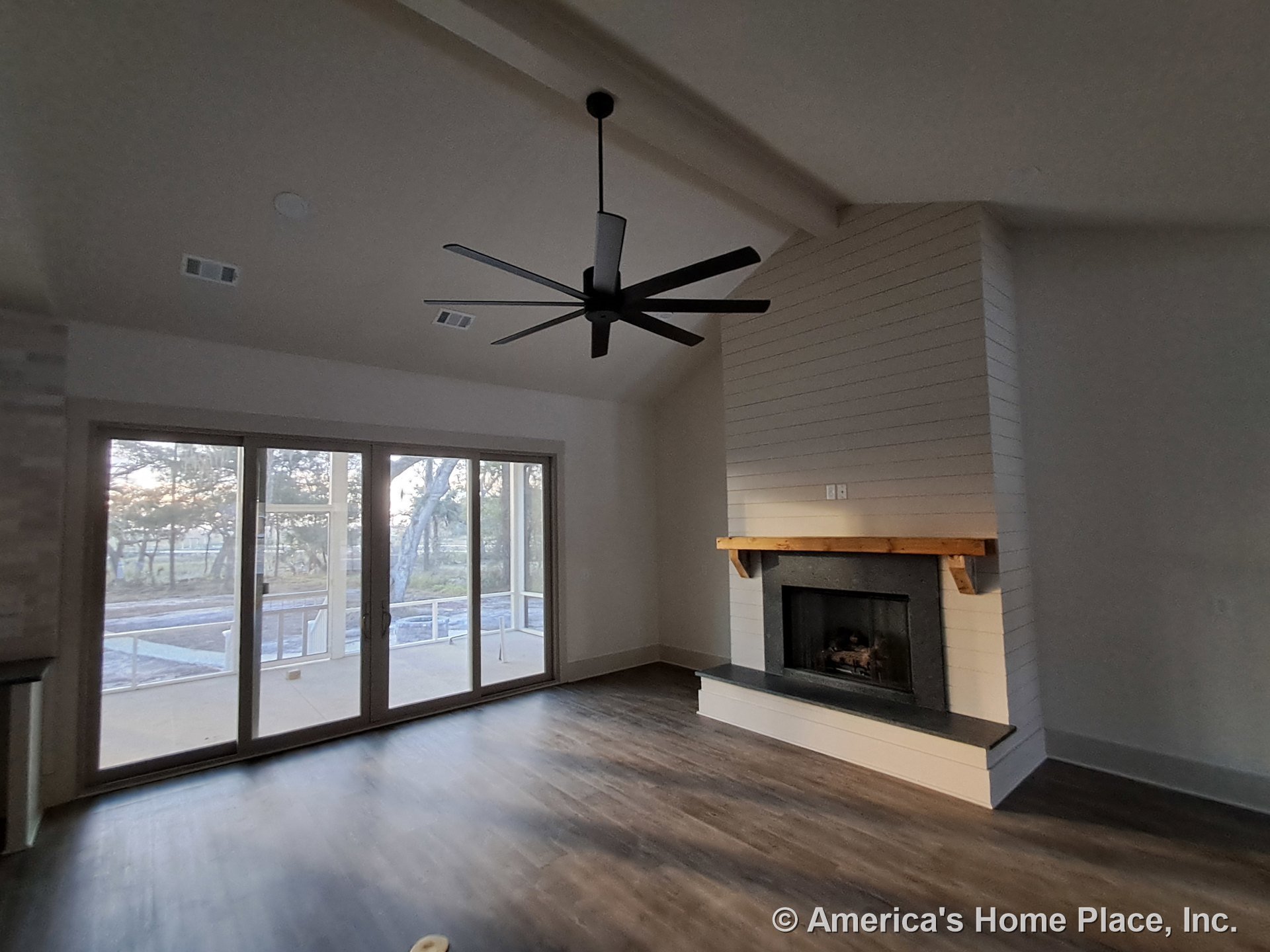 Shiplap fireplace wall with wood mantel, wide plank flooring, large sliding glass doors, vaulted ceiling with exposed beam, modern ceiling fan, and crisp window trim.