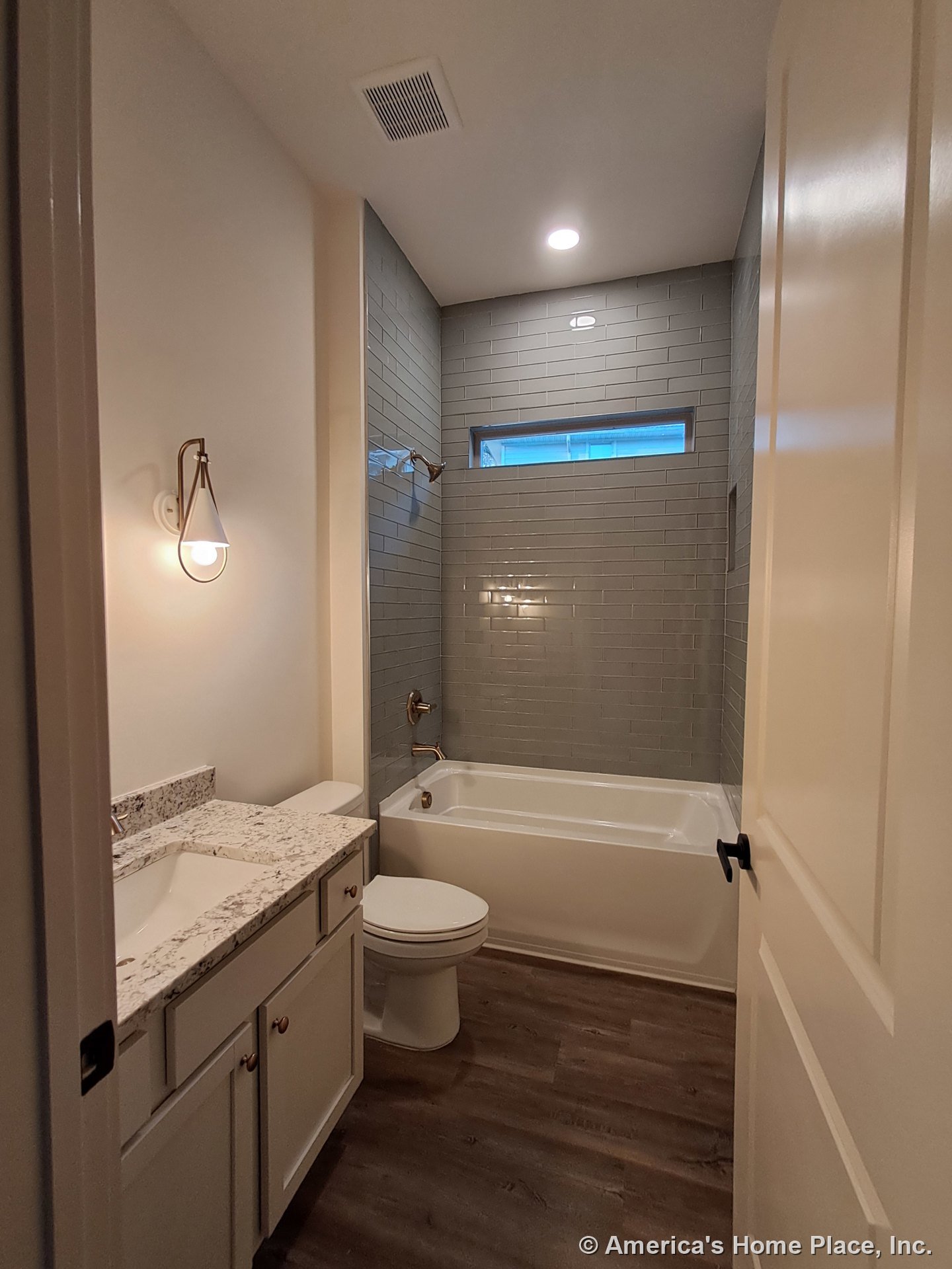Bathtub with gray subway tile surround, granite countertop vanity with single undermount sink, wood-look plank flooring, wall sconce lighting, horizontal window above tub, recessed