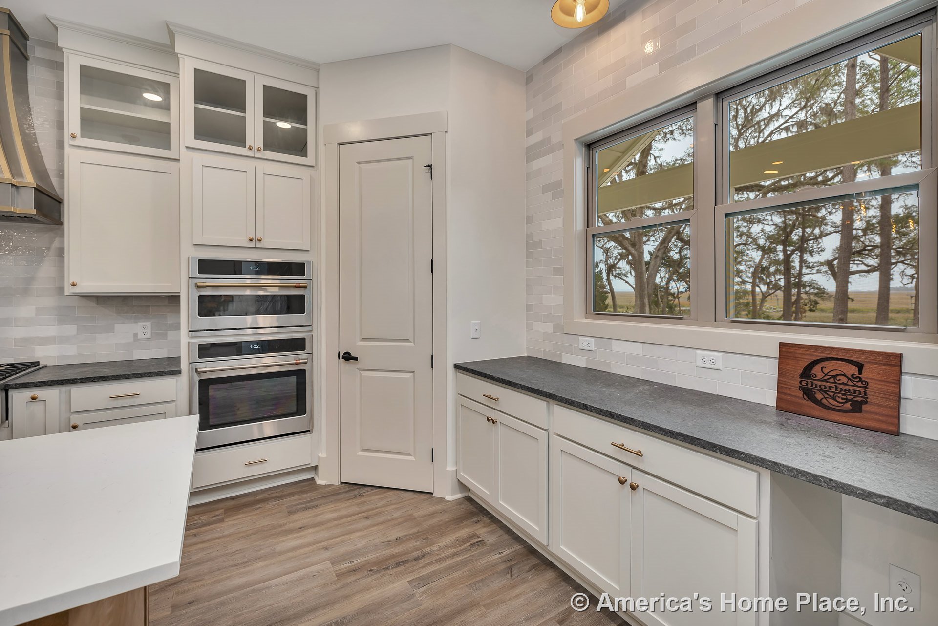 Shaker-style cabinets with brushed hardware, quartz countertops, triple stainless steel wall oven, tile backsplash, double window above sink, wood-look flooring, built-in lighting