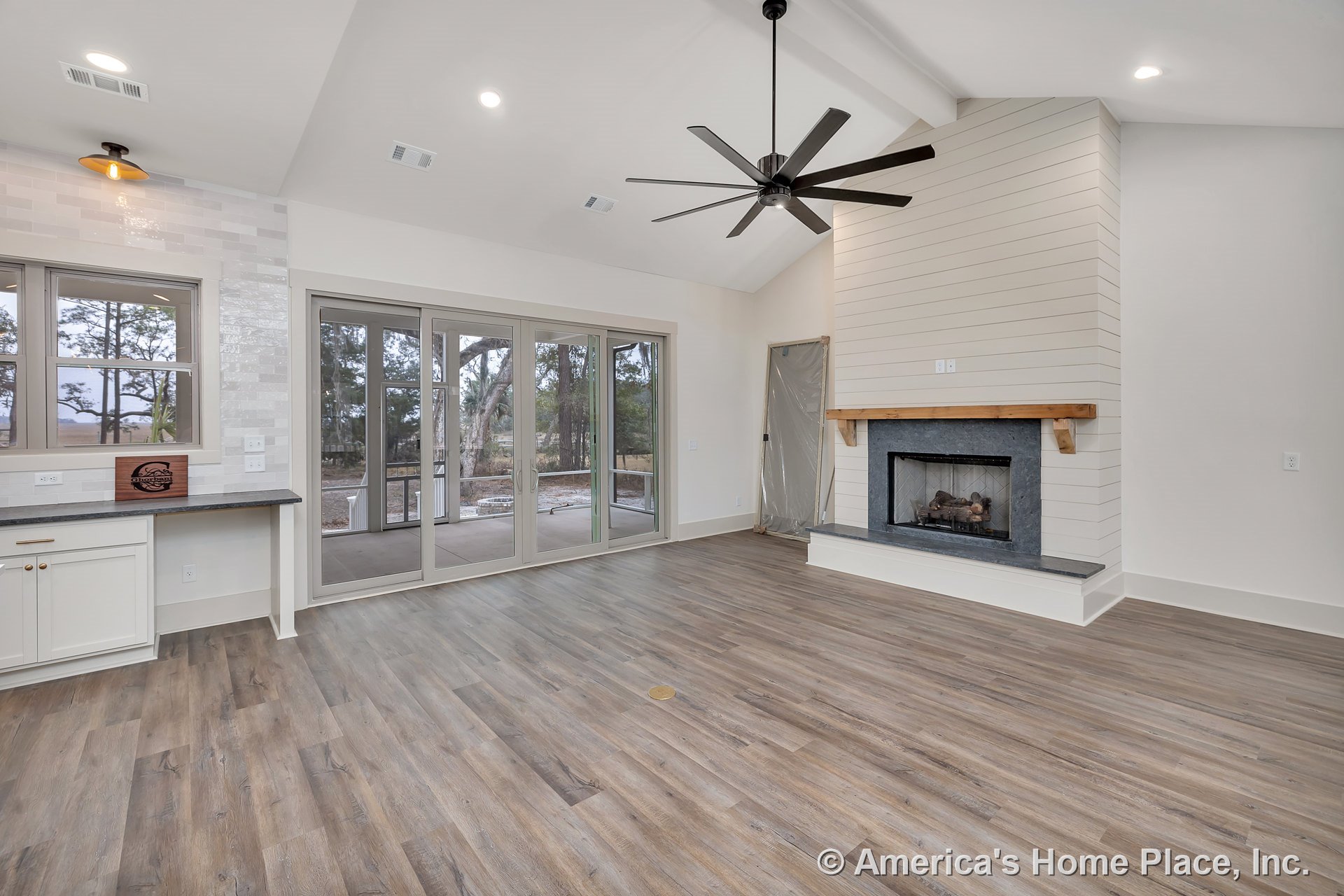 Shiplap fireplace wall with wood mantel, built-in cabinetry topped with stone countertop and tile backsplash, wide plank wood-look flooring, large sliding glass doors, ceiling fan