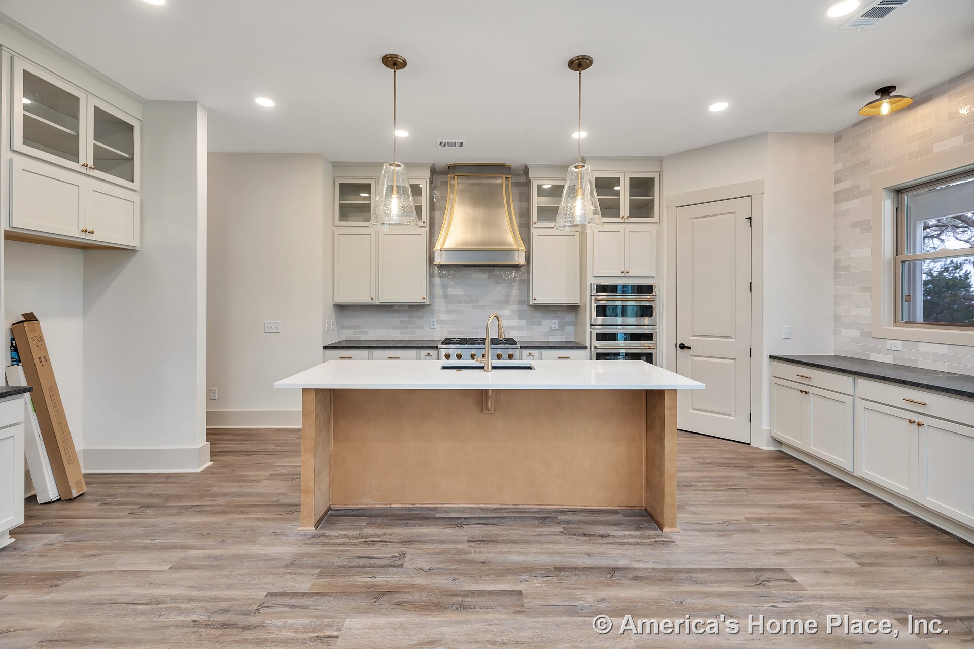 Central kitchen island with white countertop, glass pendant lights, stainless steel range hood, built-in double wall ovens, white shaker cabinets, tile backsplash, wood-look