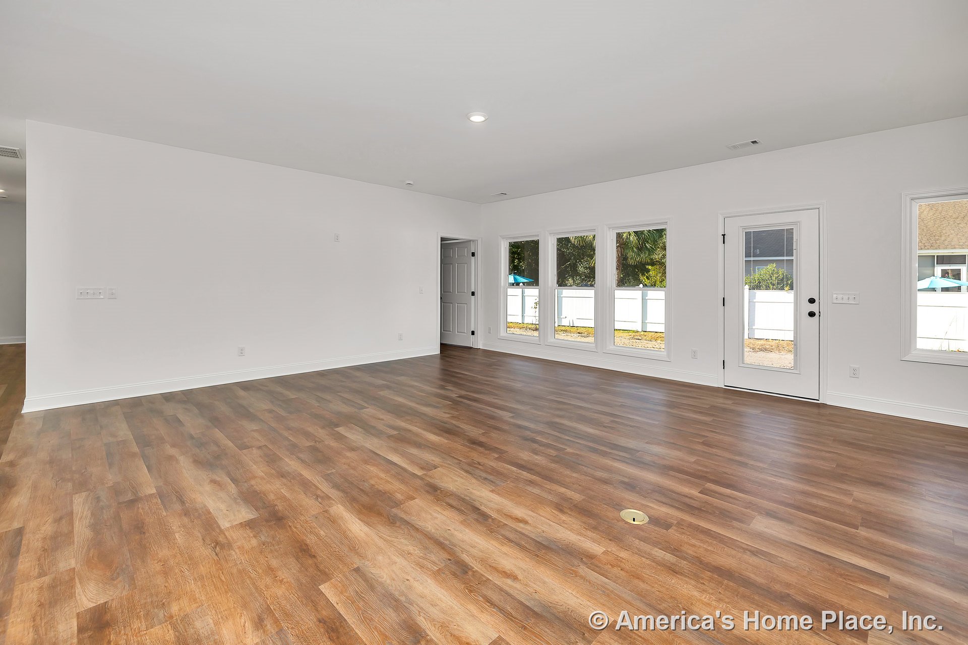 Open living area with wide plank vinyl flooring, white walls and trim, three-panel windows, glass exterior door, recessed ceiling lighting, and baseboards in a neutral color