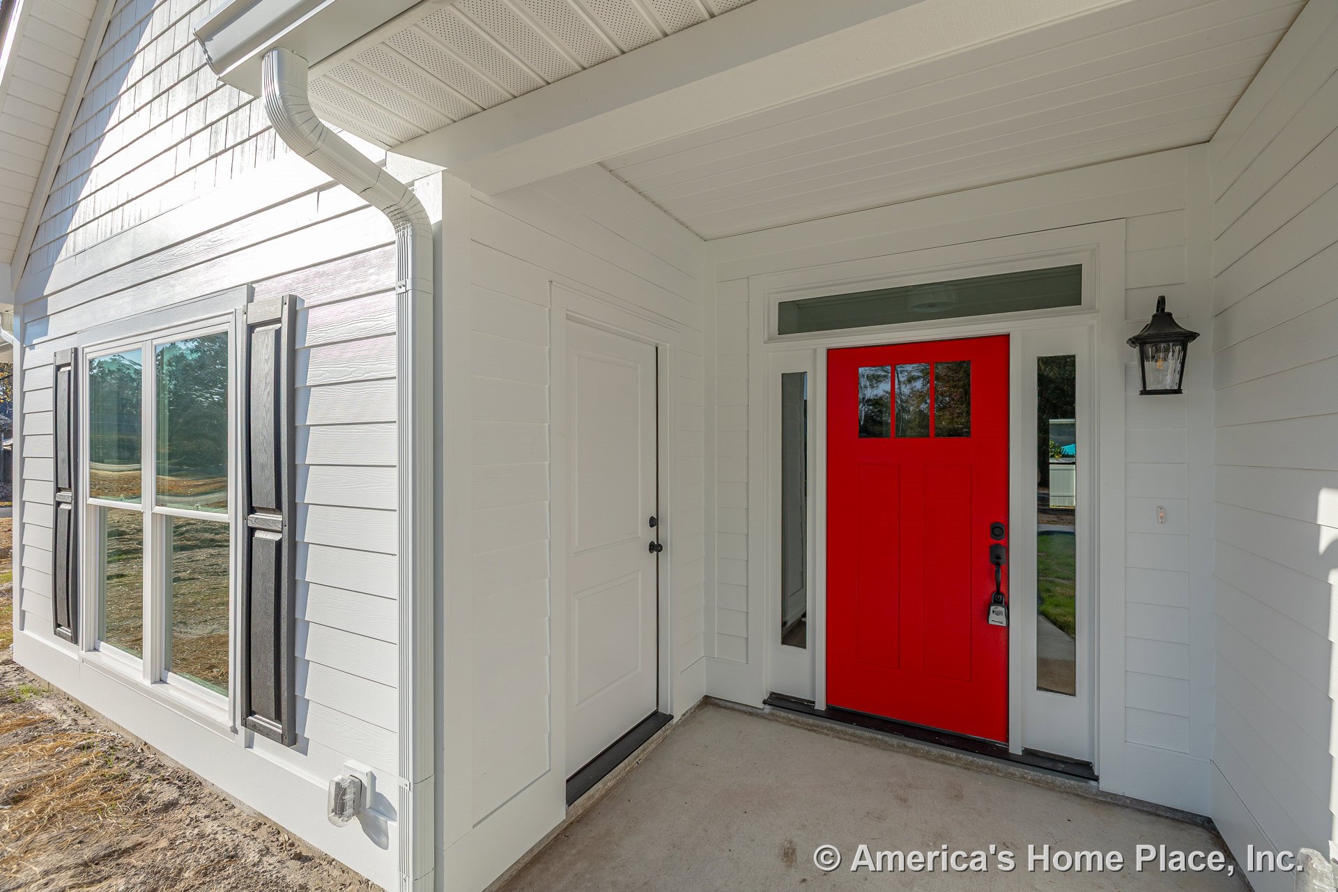Covered entry porch with white horizontal siding, bold red front door featuring glass sidelights and transom window, black exterior lantern, double window with black shutters, and