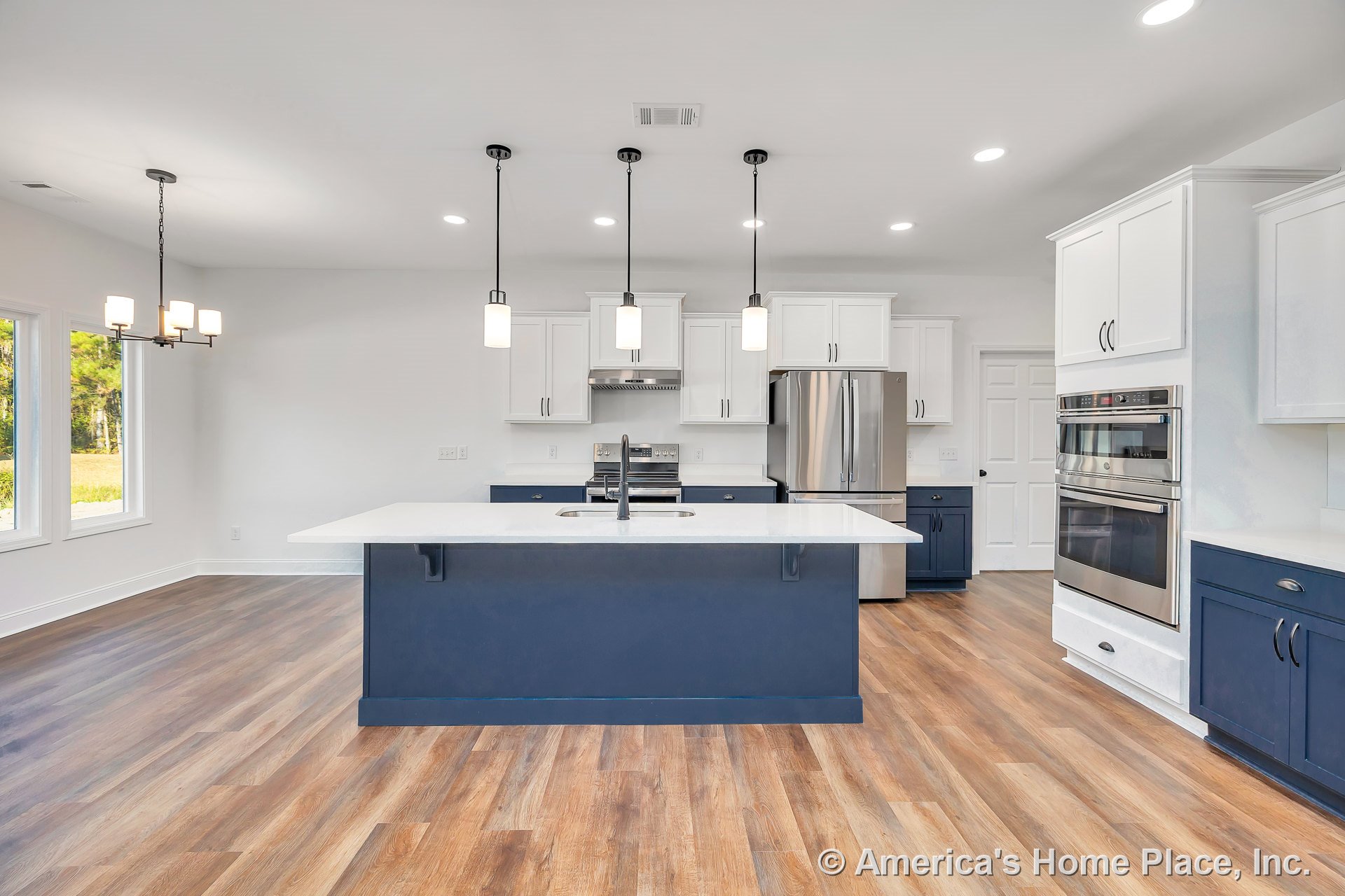 Spacious kitchen with central quartz island, blue lower and white upper cabinetry, pendant lighting, double wall oven, stainless steel refrigerator, wood-look flooring, and open