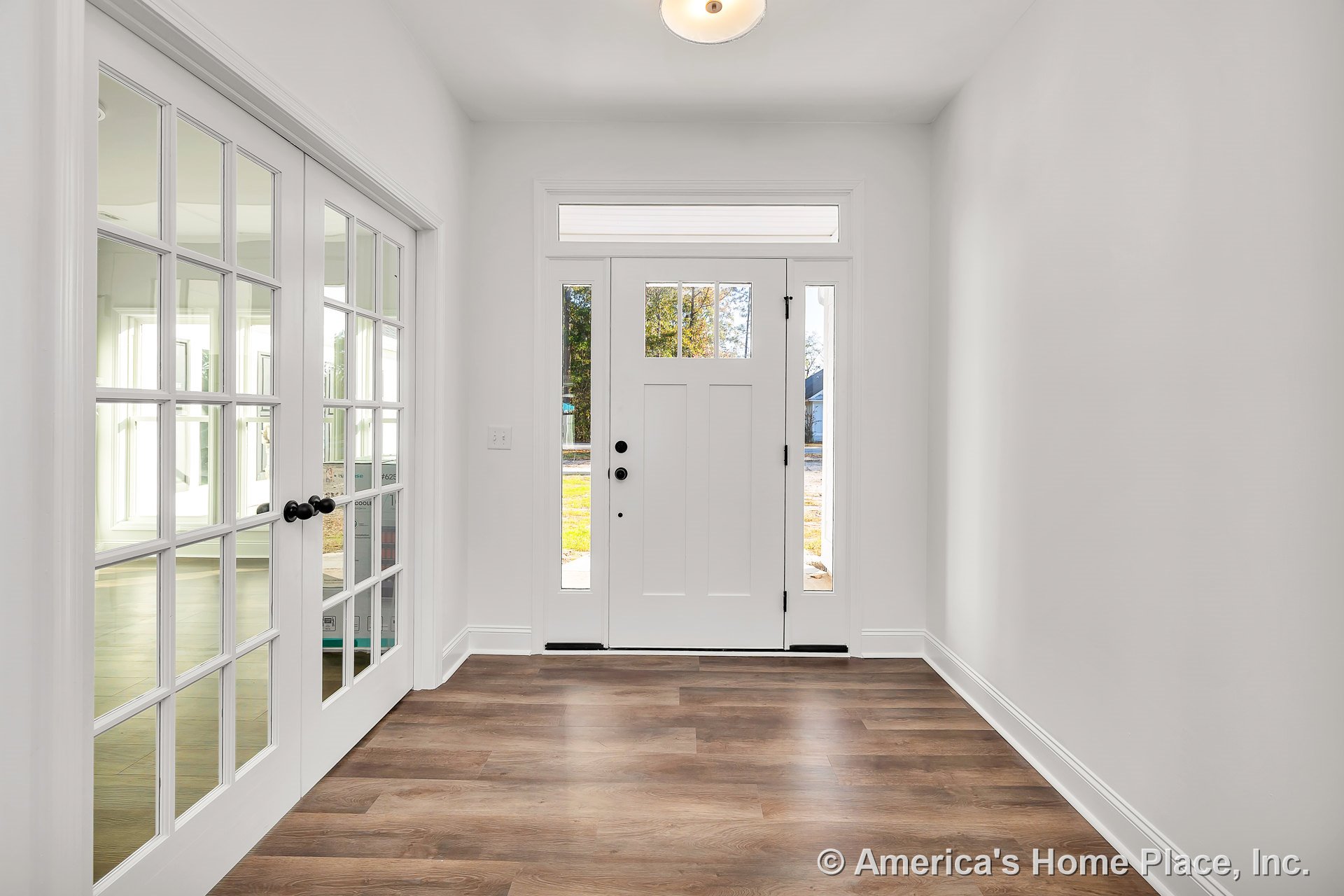 White paneled front door with sidelights and transom window, wood-look plank flooring, double glass French doors, white trim and baseboards, flush mount ceiling light, natural