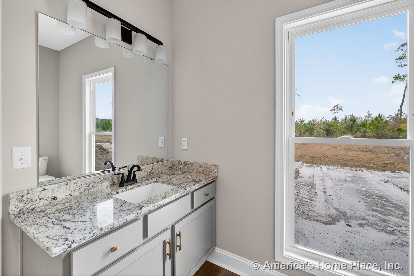 Granite vanity with undermount sink, single faucet, gray cabinetry, large wall mirror beneath four-light fixture, white-trimmed window, neutral wall color, wood flooring, and wall